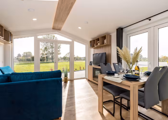 G-Spot – Moderne woonkamer met blauwe bank, eettafel, tv-meubel en grote ramen met uitzicht op groen grasland.