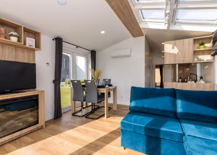 G-Spot – Moderne woonkamer met een blauw fluwelen bank, houten tafel met stoelen, tv en grote ramen naar buiten.