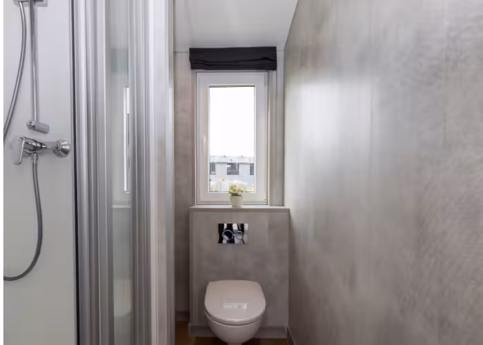 G-Spot – Kleine badkamer met hangend toilet, glazen douchecabine en raam met zwart rolgordijn.