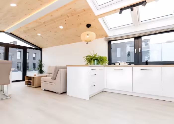 Capri – Moderne woonkamer met beige bank, witte keuken, grote ramen en houtafwerking aan het plafond.
