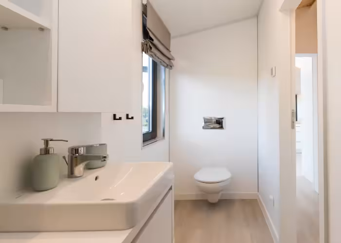 Capri – Moderne badkamer met een witte wastafel, dispenser, raam en hangend toilet.