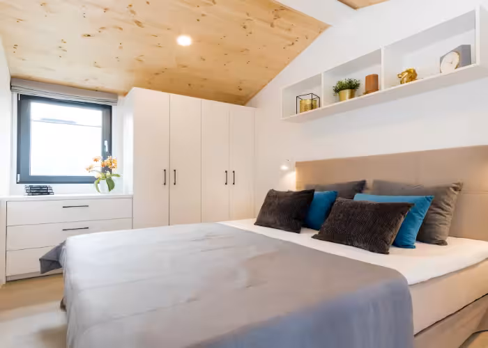 Capri – Slaapkamer met wit bed, grijze dekens, kussens in zwart en blauw, witte kast en houten plafond.
