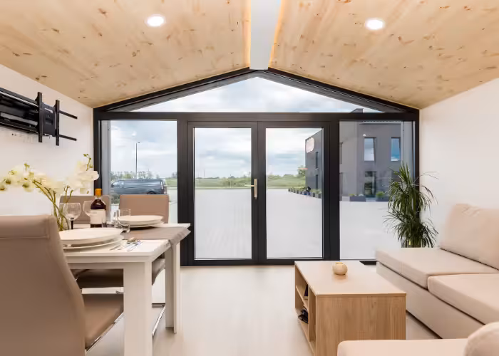 Capri – Leefruimte met houten plafond, beige bank, eethoek en grote glazen deuren naar buiten.