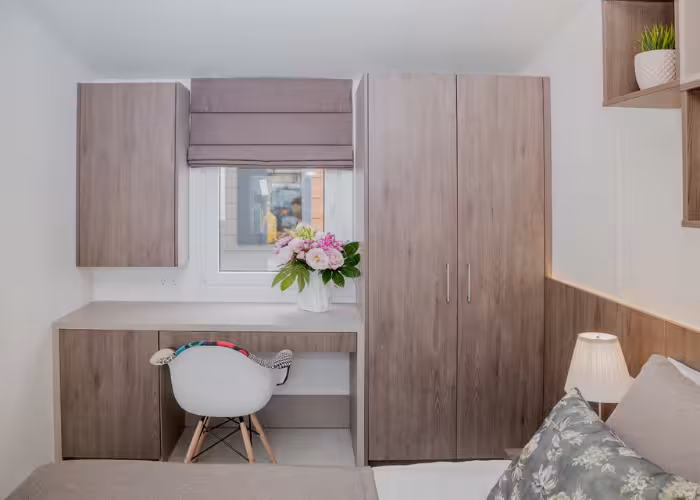 Korfu – Slaapkamer met houten kast, bureau met bloemen in vaas en een witte stoel met kleurrijke armleuningen.