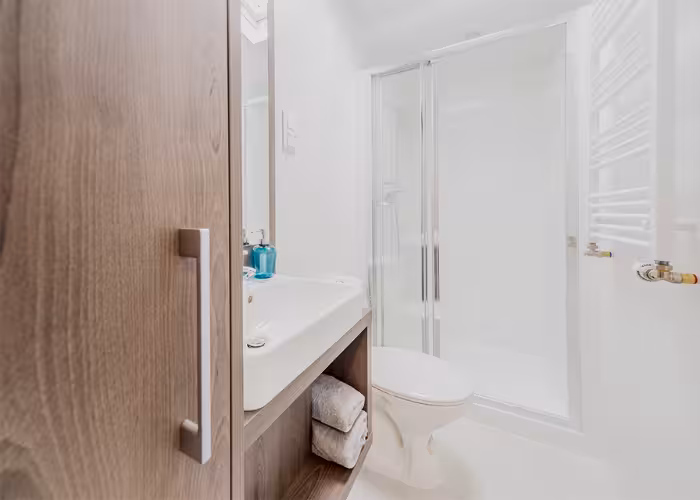 Korfu – Kleine moderne badkamer met houten kast, witte wastafel, toilet en glazen douchewand.