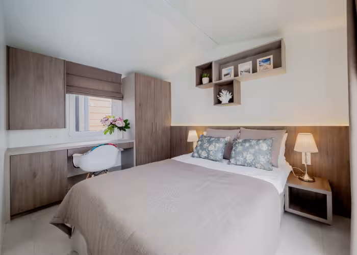 Korfu – Slaapkamer met tweepersoonsbed, nachtkastjes, bureau, witte stoel en decoratieve planken aan de muur.
