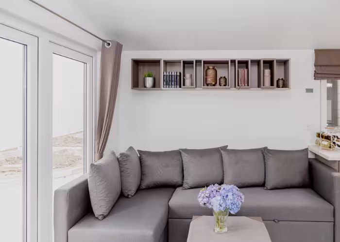 Korfu – Lichte woonkamer met grijze hoekbank, bloemenvaas op tafel en zwevende plank met decoratie en boeken.