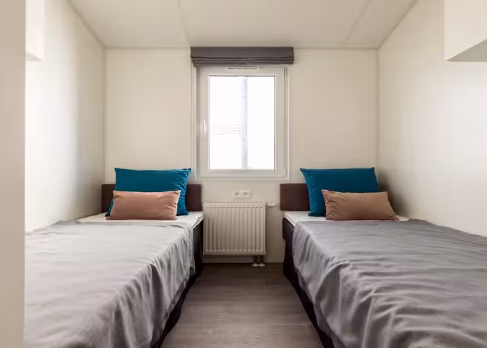 Texel – Twee eenpersoonsbedden met grijze bedspreien en kussens naast een raam in een minimalistische kamer.