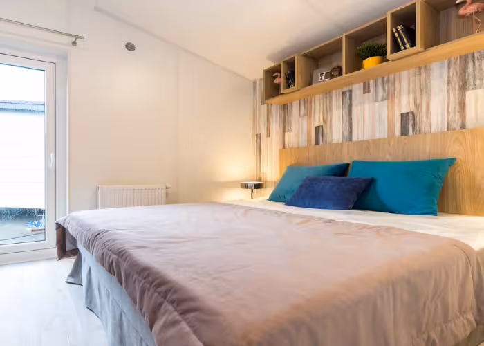 Murano – Moderne slaapkamer met groot tweepersoonsbed, blauw en turquoise kussen, houten wanddecoratie en open kast.
