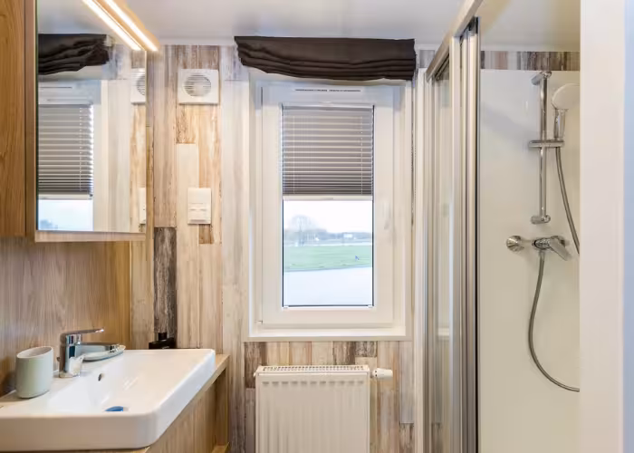 Murano – Badkamer met wastafel, spiegel, raam, radiator en een douchecabine met douchekop en slang.
