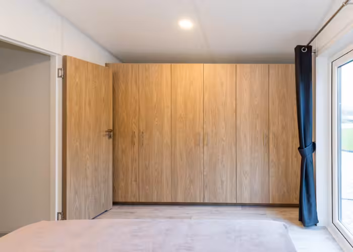 Murano – Slaapkamer met ingebouwde houten kast, open deur en zwart gordijn bij glazen raam.