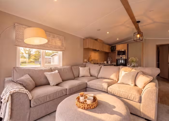 Arlington – Lichte beige hoekbank in woonkamer met ramen, lamp, ronde poef en open keuken op de achtergrond.
