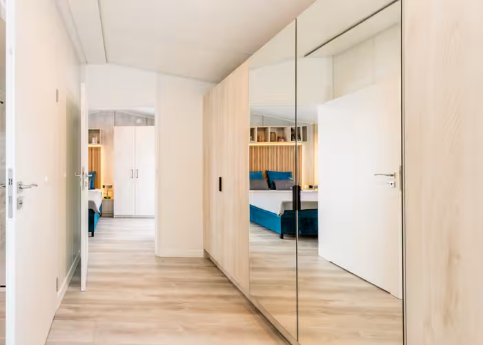 Chios – Moderne gang met licht houten vloer en kasten met spiegels die een slaapkamer met een bed weerspiegelen.