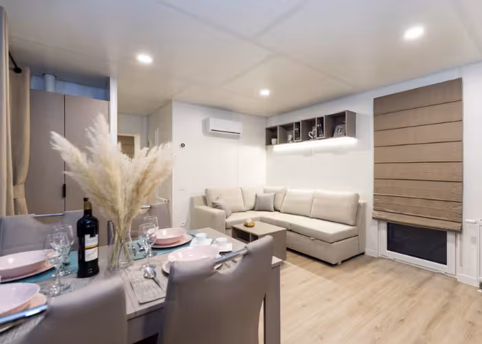 Joli – Moderne woonkamer met beige hoekbank, eettafel met glazen en een fles wijn, en houten vloer.