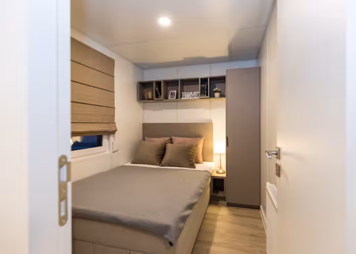 Joli – Knusse slaapkamer met tweepersoonsbed, bruine gordijnen, kast en zichtbare boekenplank met decoratie.