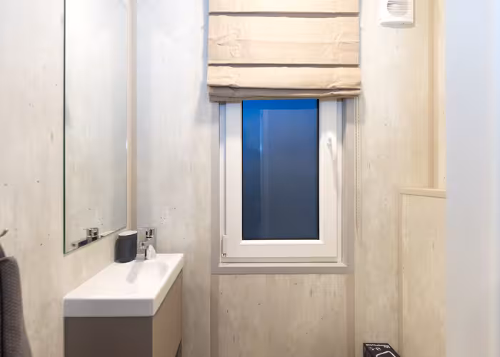 Joli – Kleine badkamer met wastafel, spiegel, raam met beige rolgordijn en lichte houten muren.