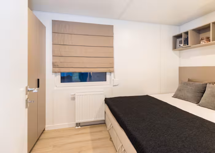 Joli – Kleine slaapkamer met dubbel bed, beige jaloezie, kast en houten vloer.