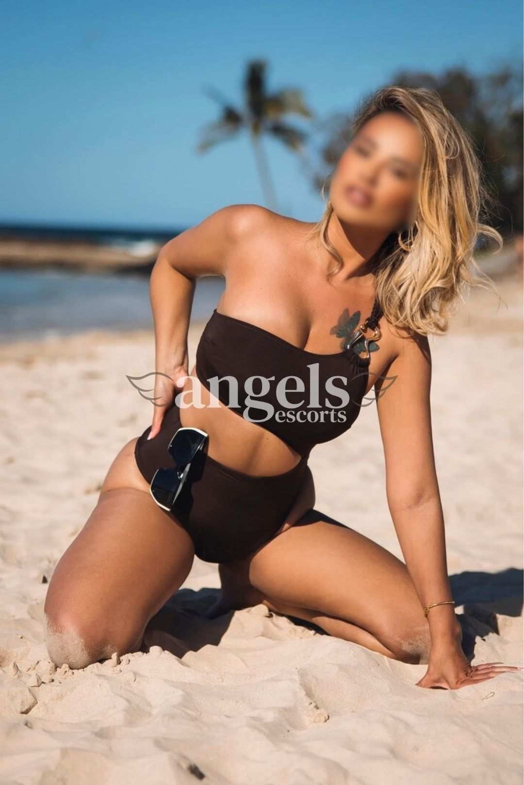 Irina kneeling on the sand in a brown bikini with bright sunlight and ocean behind her.