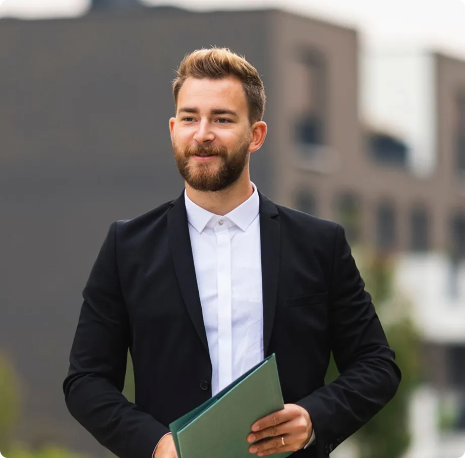 Confident young man in a black suit holding a green folder outdoors with a blurred building background.