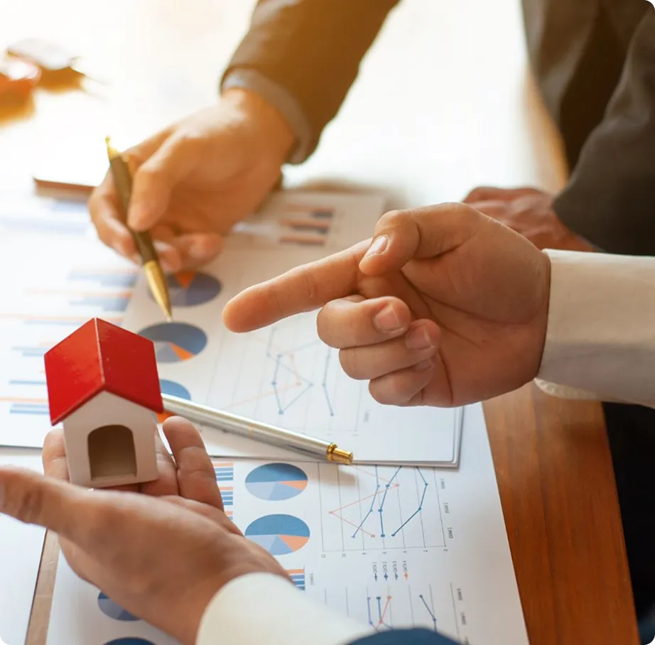 Hands holding a small house model and pointing at financial charts with graphs and pie charts on a table.