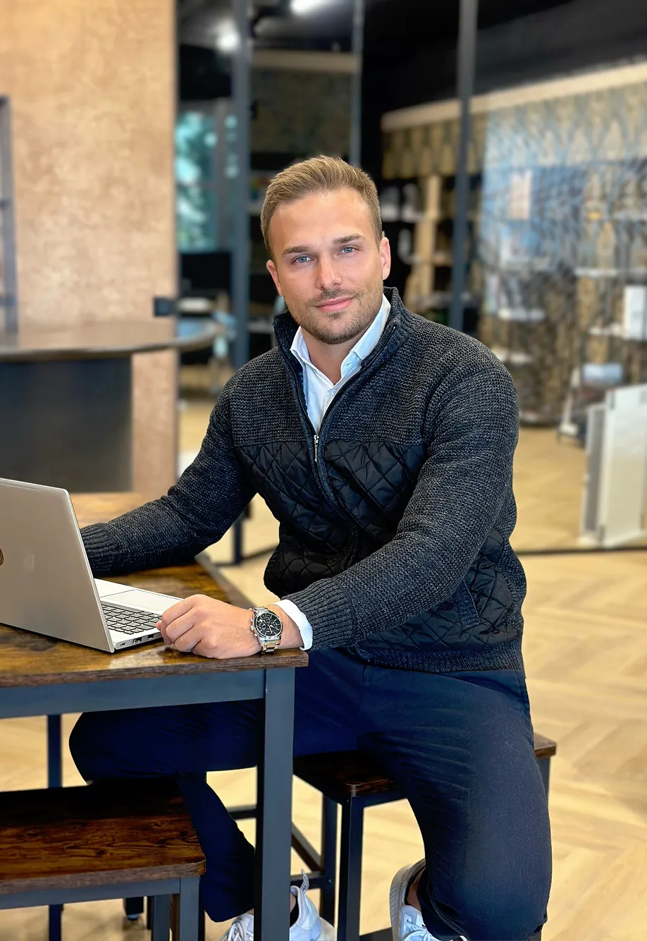 Man wearing a black quilted jacket sitting at a wooden table working on a laptop in a modern office.