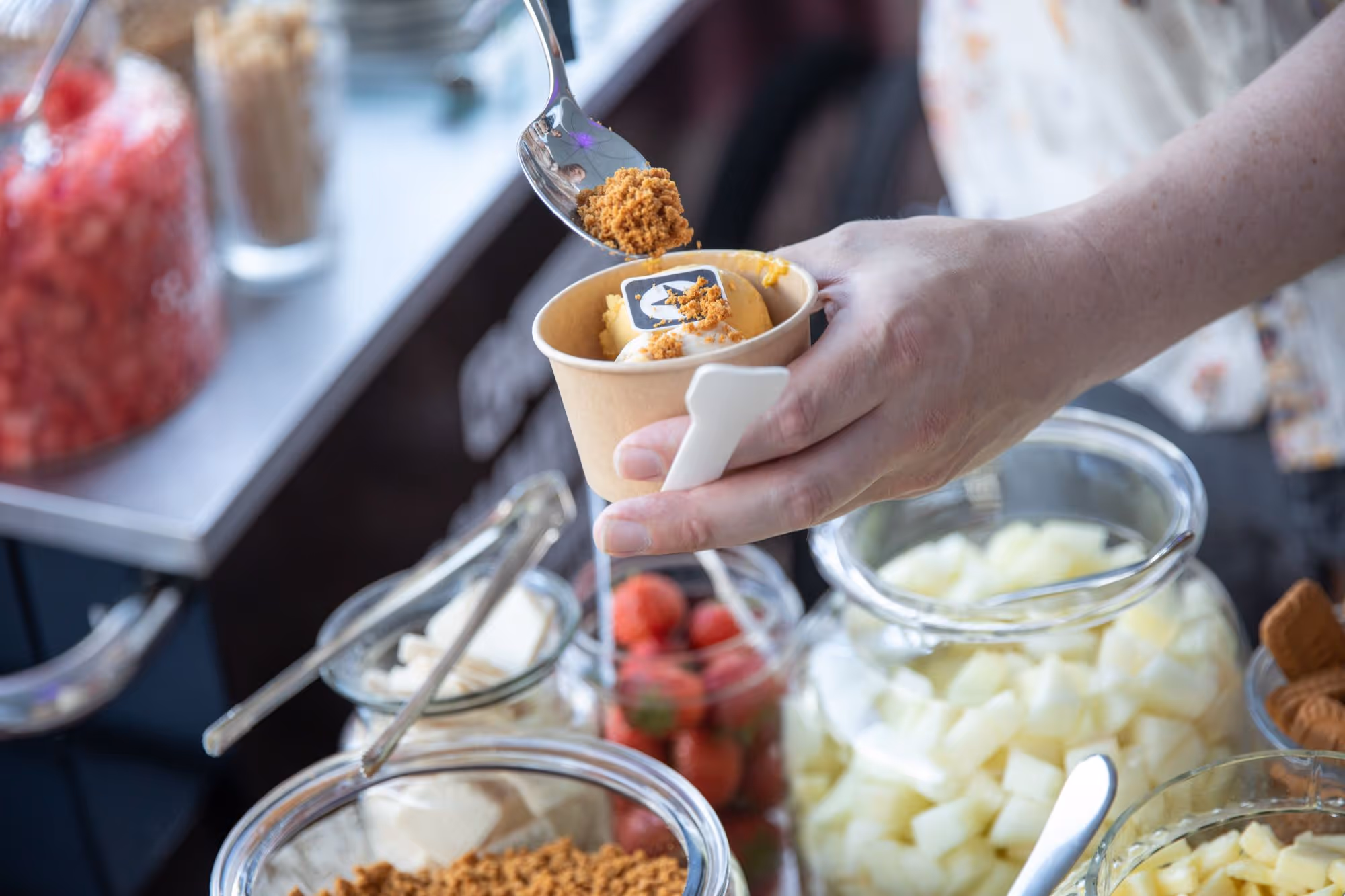 Hand hält einen Pappbecher mit Eis und streut braune Streusel darauf, im Hintergrund verschiedene Gläser mit Obst und Zutaten für das Signature-Eis beim Congstar Sommerfest 2025.