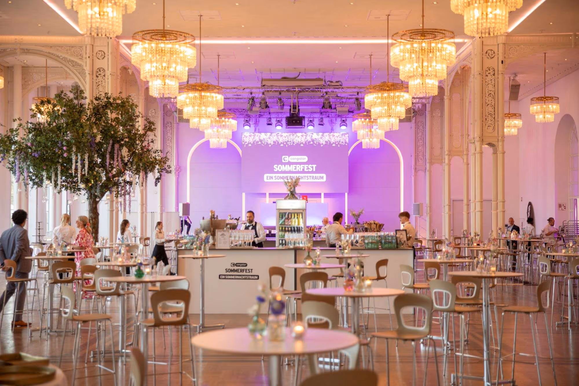 Großer eleganter Festsaal mit Kronleuchtern, hohen Decken und Tischen, an denen Menschen beim Sommerfest von Congstar mit dem Motto 'Ein Sommernachtstraum' stehen.