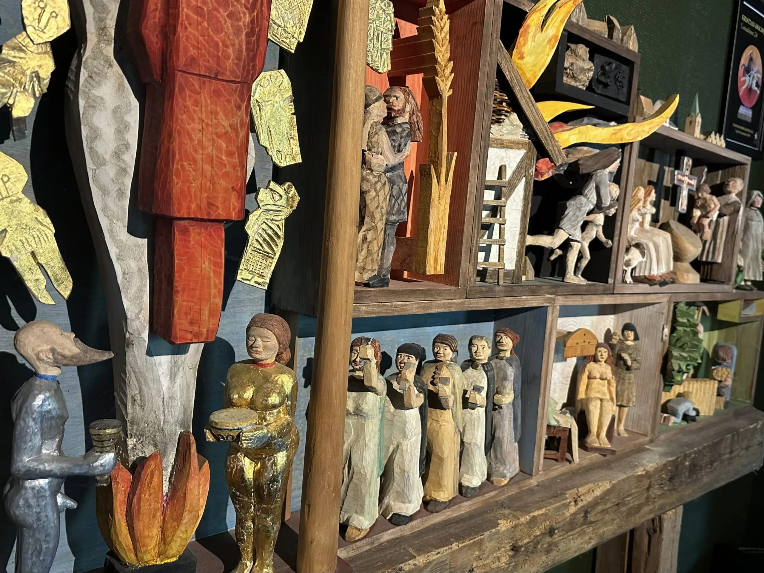 Träskulpturer av olika mänskliga figurer i små fack, inklusive en guldfärgad figur med trumma och en grå figur med näbb, samt detaljer som eld och byggnader.