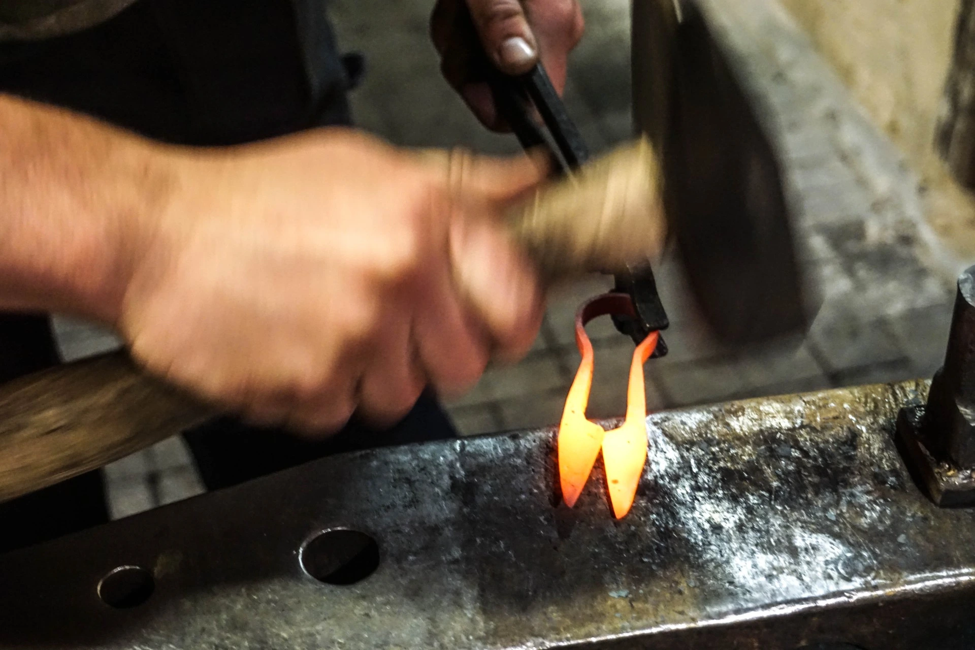 Hantverkare smider upphettade, glödande metallstycken på ett städ med hammare och tång.