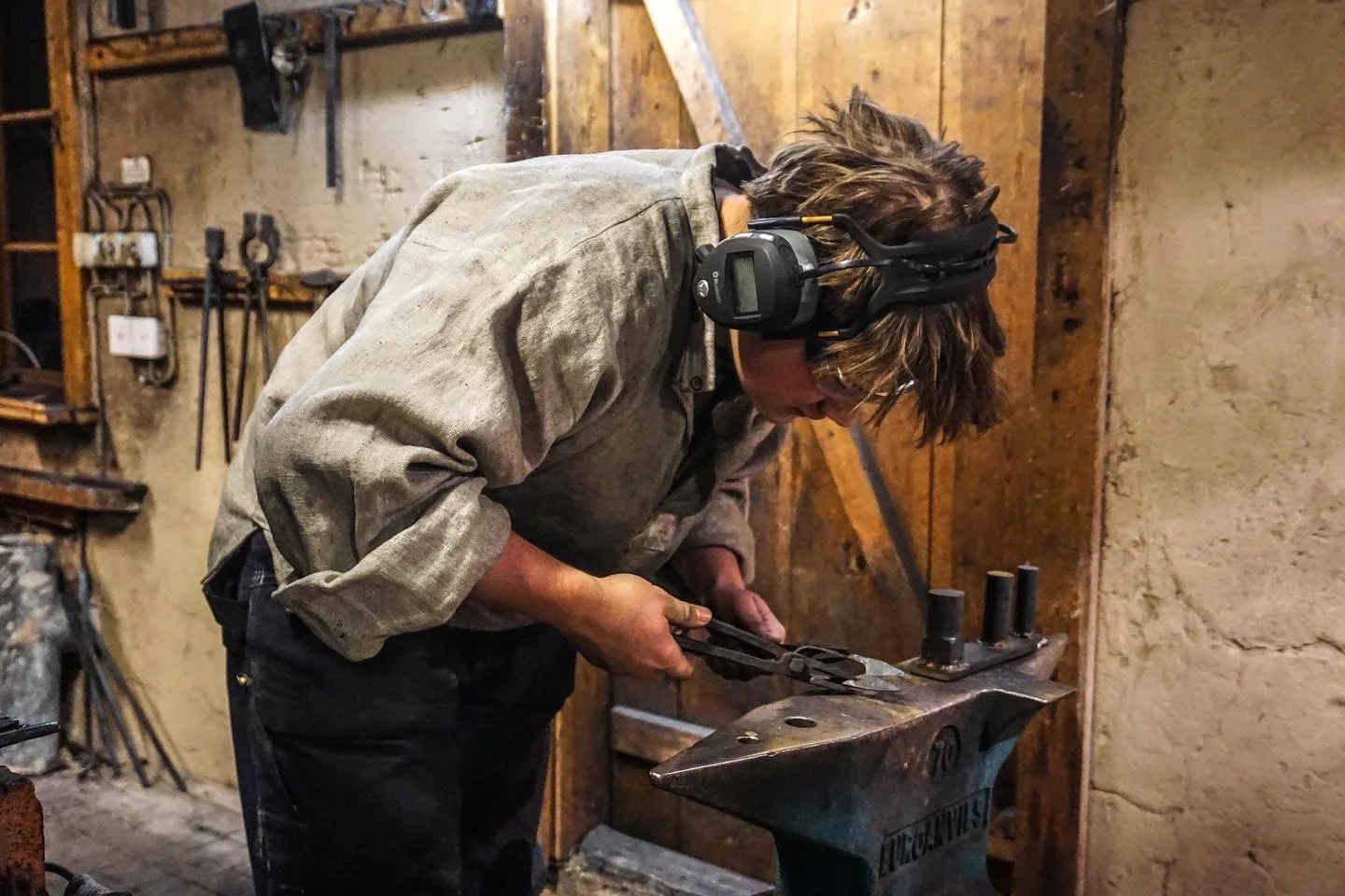En smed böjer sig över ett städ och arbetar med tång i en verkstad, iförd skyddshörselkåpor.
