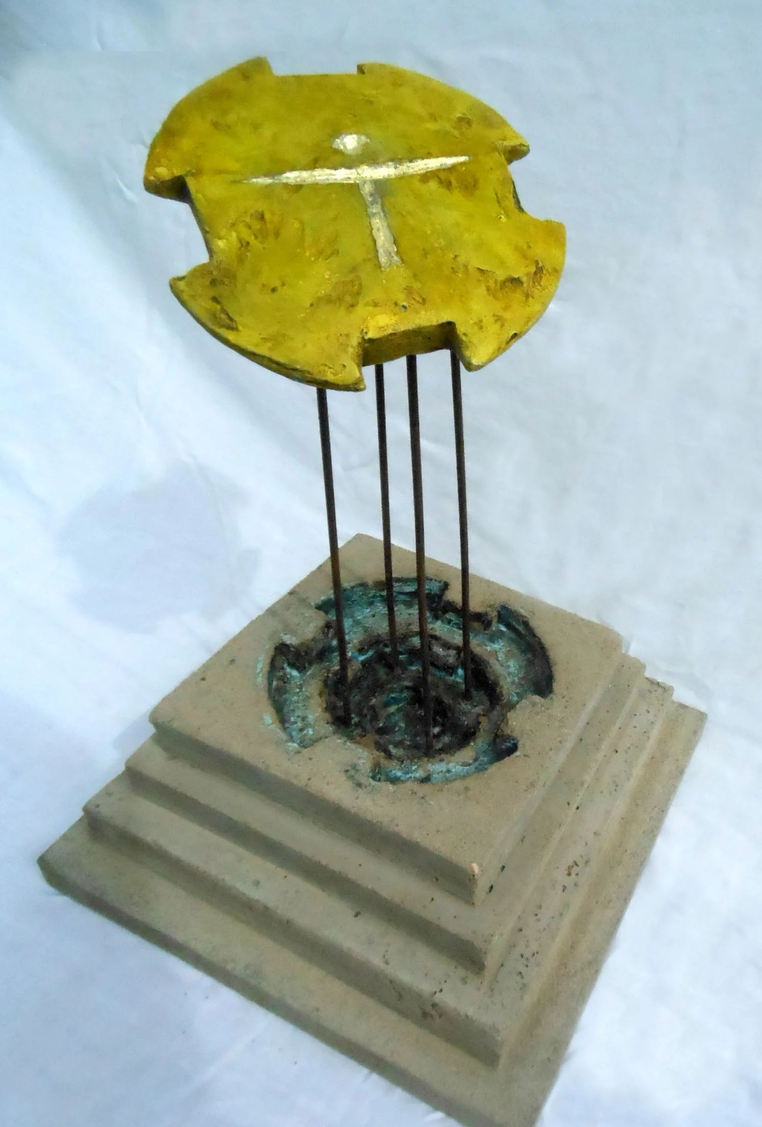 Konstverk med en gul cirkulär skiva prydd med ett vitt T-format mönster, upphöjd på en fyrstegsbas av betong med fyra smala metallstavar.