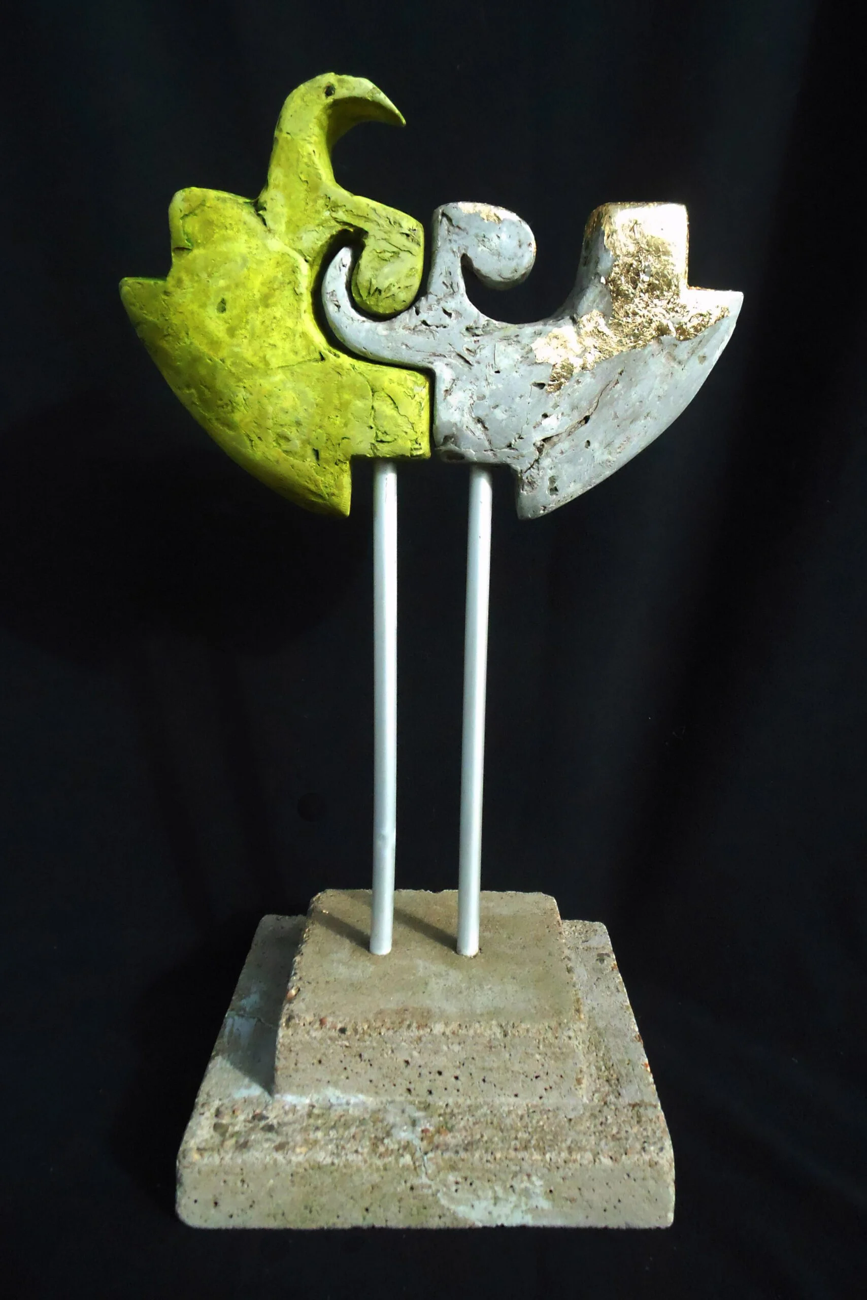 Abstrakt skulptur i form av två sammanfogade fåglar, en gul och en vit, monterade på två vita stänger på en betongbas.
