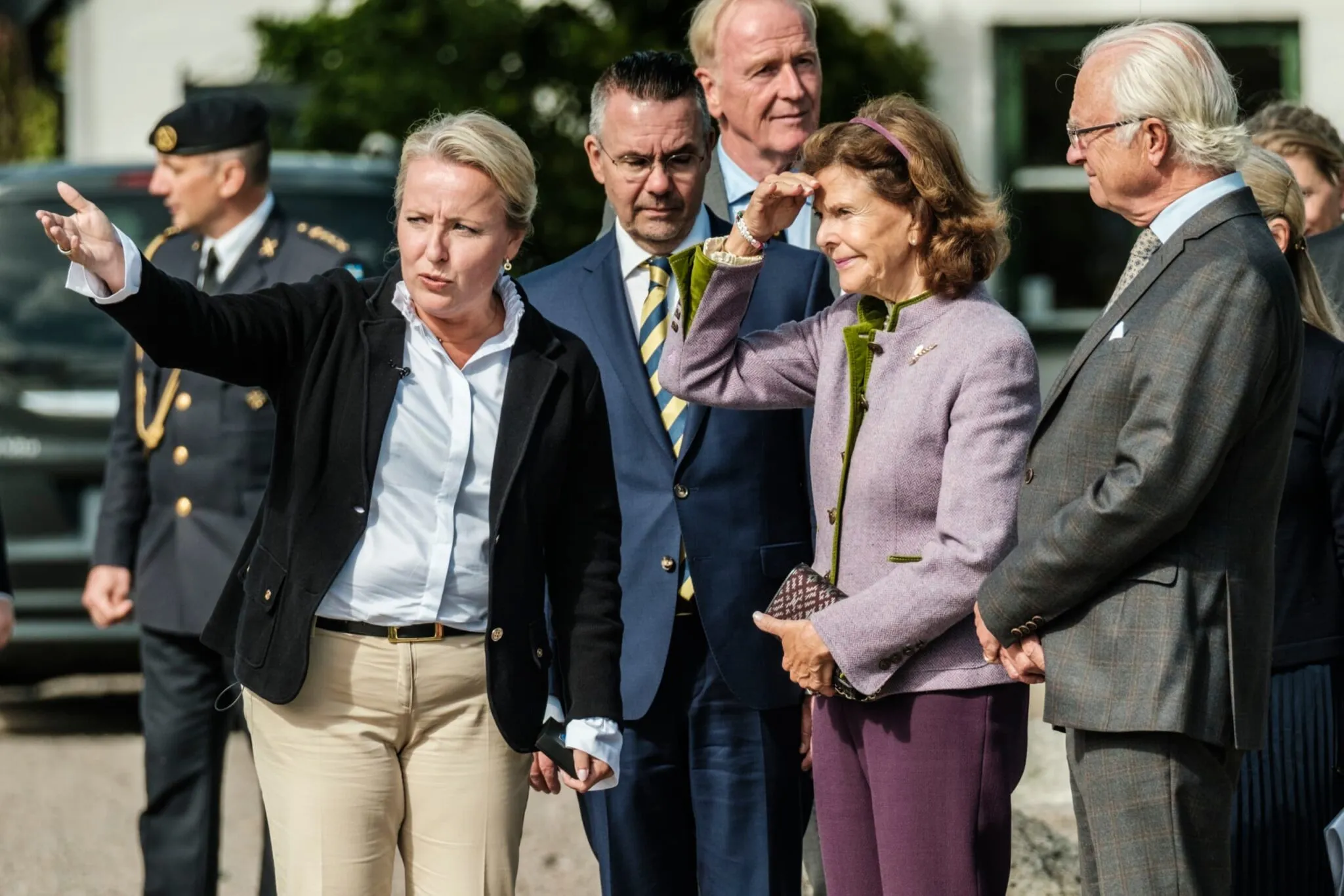 En kvinna i svart kavaj pekar åt vänster medan en grupp människor inklusive en äldre man och kvinna i lila kavaj tittar åt samma håll utomhus.