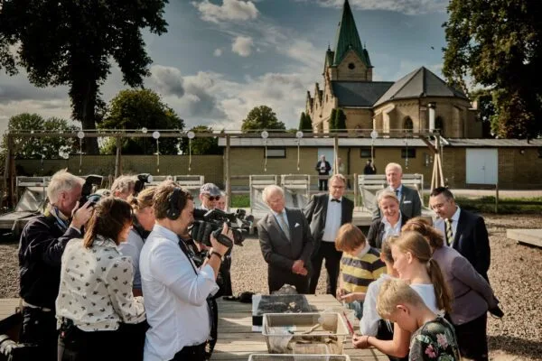 Grupp av vuxna och barn samlade utomhus runt ett bord med skålar, med en kyrka i bakgrunden under en molnig himmel.