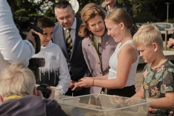 En grupp barn och två vuxna, inklusive en kvinna i lila jacka och en man i kostym, tittar tillsammans på något i en burk eller låda under en utomhusbelysning.