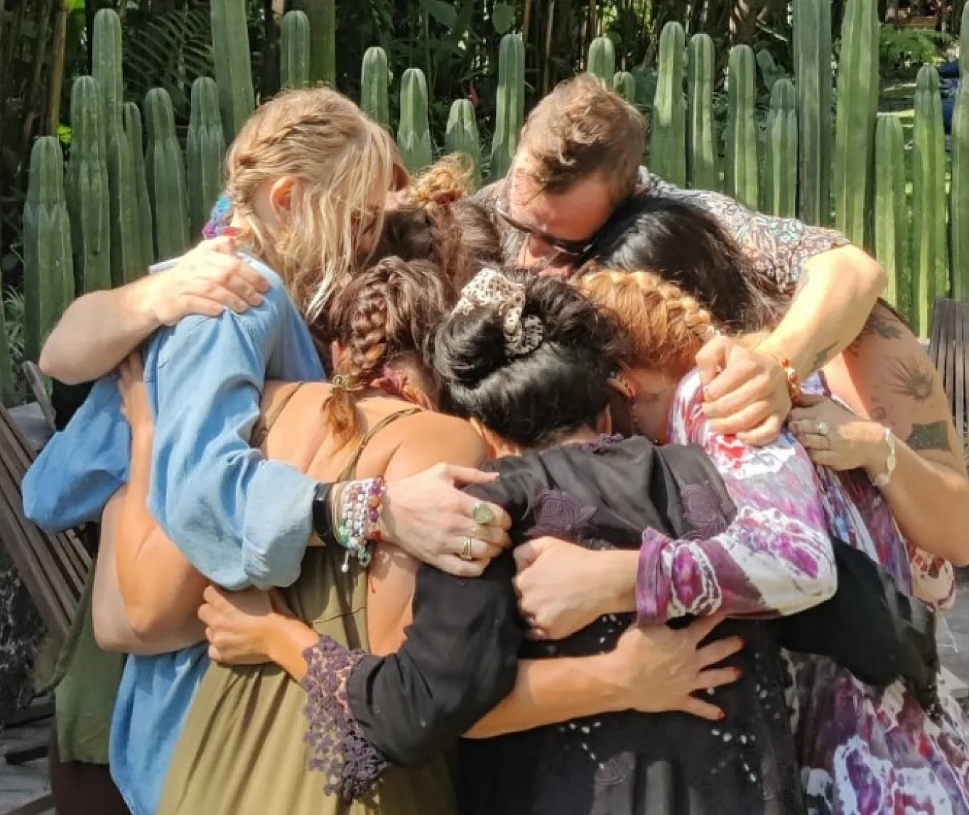Group of seven people embracing in a close group hug outdoors with tall cacti in the background.