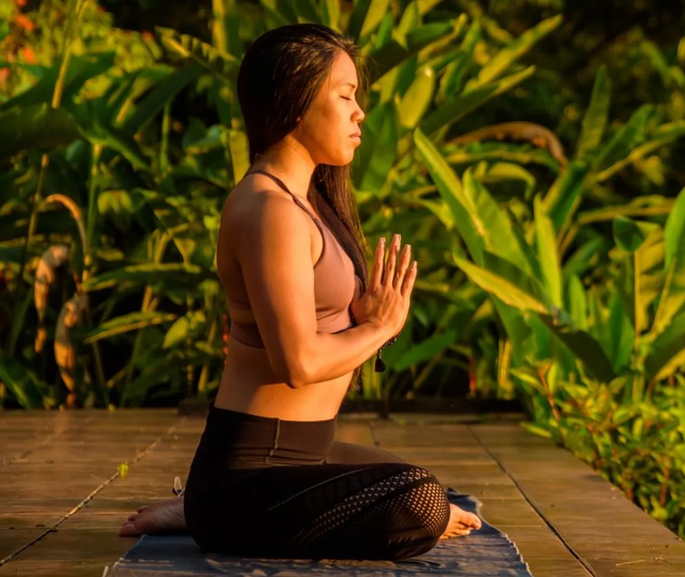 Woman meditating outdoors on a yoga mat surrounded by lush green plants during golden hour.