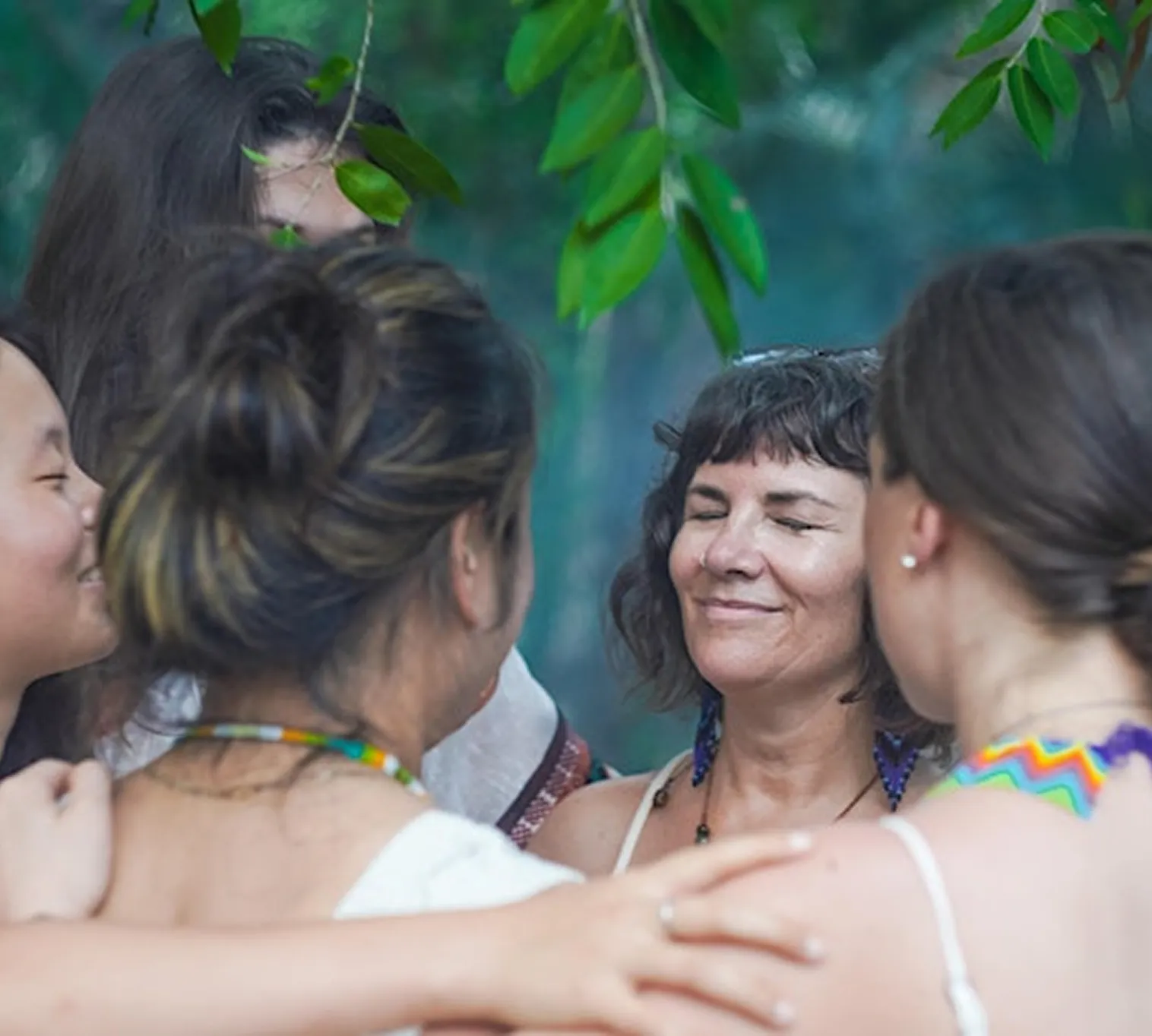 Group of five women standing close together outdoors, smiling and embracing under green leaves.