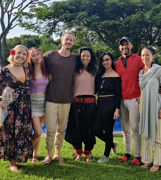 Group of seven adults standing together outdoors on green grass with trees and a blue pool in the background, smiling and posing for the photo.