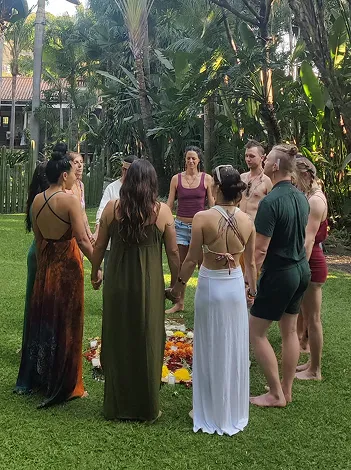 Group of barefoot people standing in a circle on grass, holding hands around a colorful flower arrangement outdoors.