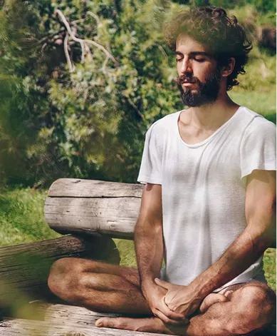 Bearded man wearing a white t-shirt sitting cross-legged on wooden steps outdoors with eyes closed, meditating.