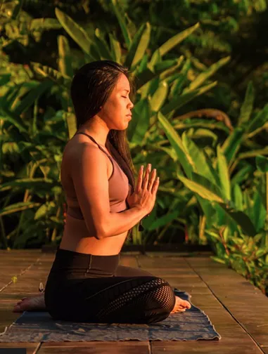 Woman meditating in a seated yoga pose on a mat outdoors with green plants in the background.