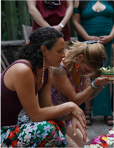 Two women sitting closely, one with floral pants and the other holding a decorative bowl with flowers, both involved in a focused, ceremonial moment.