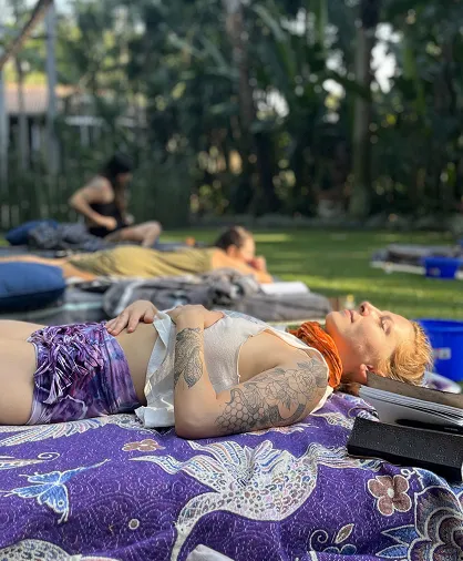 Young woman with a floral tattoo lying on a purple patterned blanket in a sunny garden, with other people relaxing in the background.