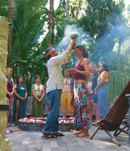 Man performing a ritual by sprinkling smoke over a barefoot woman standing with eyes closed, surrounded by people in colorful clothing in a lush outdoor setting.