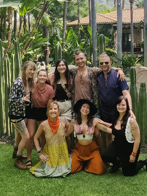 Group of eight smiling young adults posing outdoors on grass with tall cacti and tropical plants in the background.