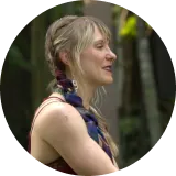 Profile of a smiling woman with braided hair, wearing a sleeveless top, outdoors with blurred greenery in the background.