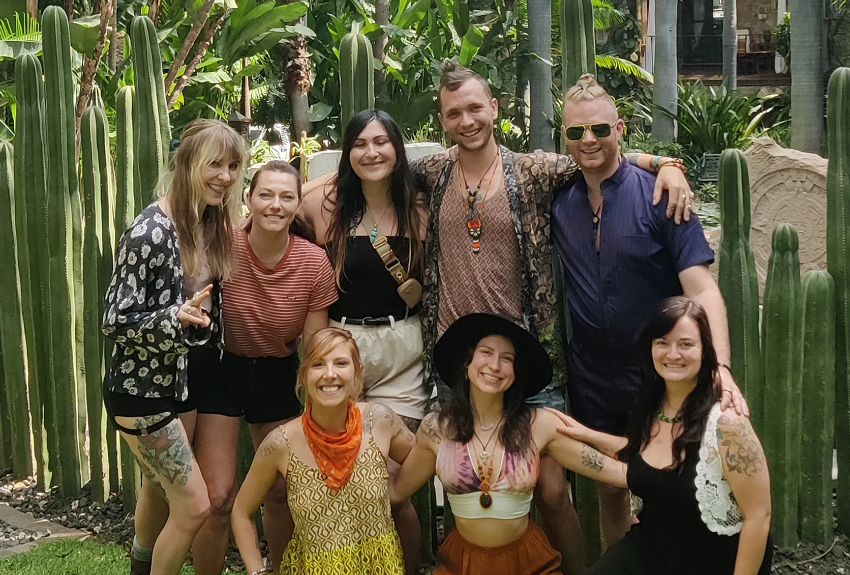 Group of eight smiling people posing outdoors surrounded by tall green cacti in a garden setting.
