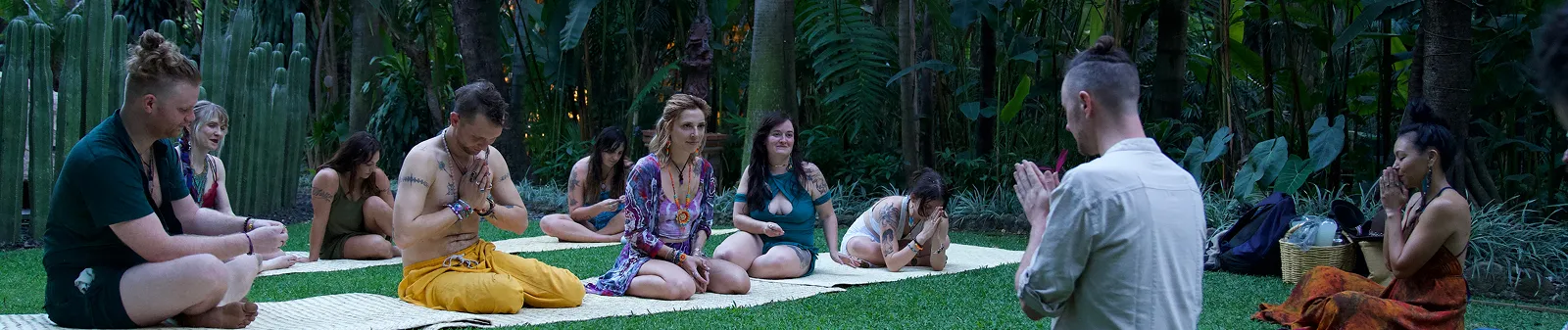 Group of people sitting on mats in meditation or prayer pose outdoors surrounded by lush greenery.