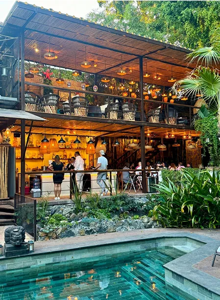 Two-story outdoor dining area with warm hanging lights, surrounded by lush greenery and a swimming pool in the foreground.